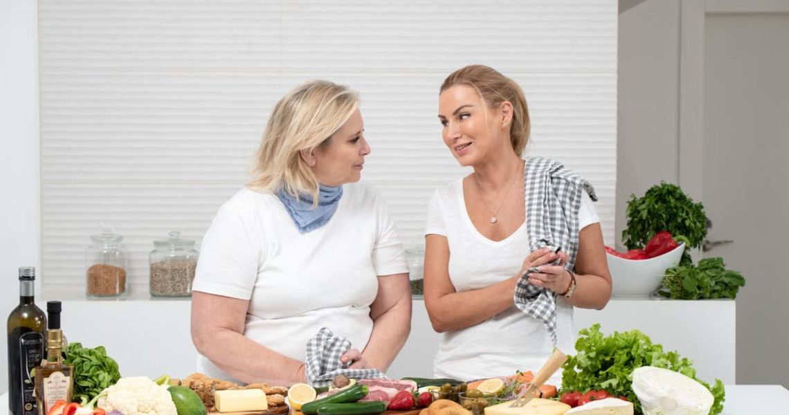 Two women in a kitchen preparing fresh vegetables and cheese with olive oil and spices.