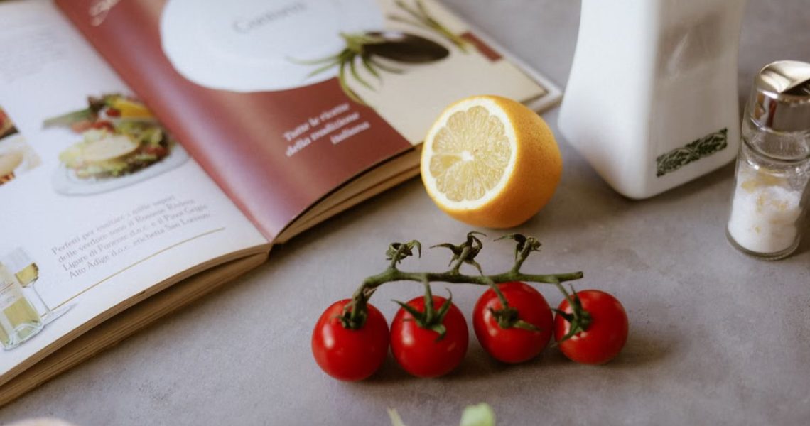 Flat lay of cooking ingredients including tomatoes and a lemon with a cookbook.
